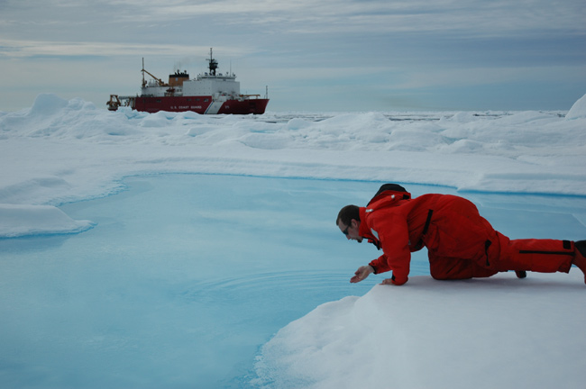 Jeremy Potter sipping water from a sea ice melt pond with the US Coast Guard Icebreaker Healy in the background